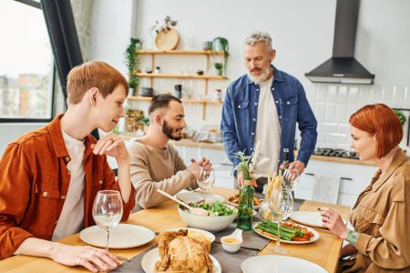bearded man standing with bottle of wine near son with gay partner during family dinner in kitchen