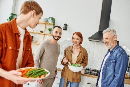 blurred gay man holding delicious asparagus near boyfriend and smiling family in kitchen
