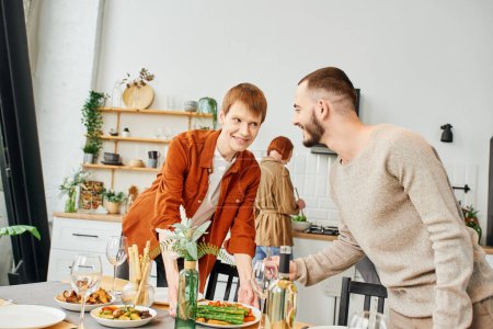 joyful gay couple looking at each other while setting table with family dinner in kitchen 