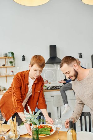 redhead gay man serving grilled asparagus near boyfriend in modern kitchen