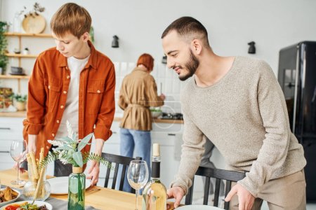 young gay couple setting family supper near parents on blurred background in kitchen