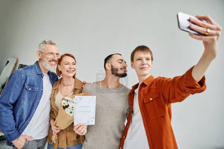 redhead man taking selfie with gay partner and happy parents holding marriage invitation and flowers
