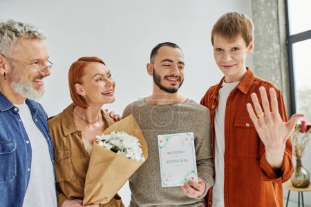 cheerful gay man showing wedding ring near boyfriend with invitation card and happy parents with flowers