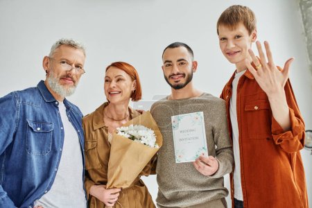 happy gay man showing wedding ring near parents with flowers and boyfriend with wedding invitation