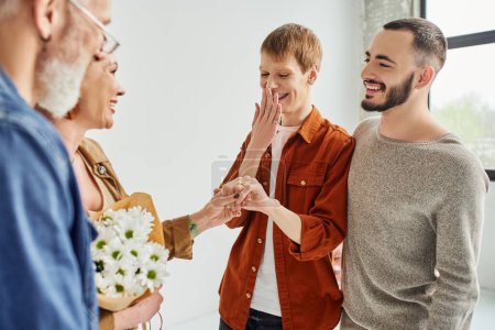 happy gay man covering mouth while showing wedding ring to happy mother with flowers at home