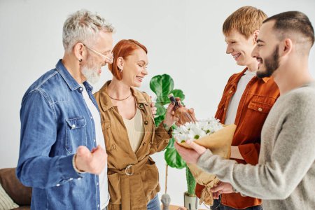 fröhliches homosexuelles Paar schenkt Blumen und Weinflasche, während es glückliche Eltern zu Hause besucht