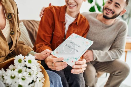 happy gay man holding wedding invitation near boyfriend and mother with flowers at home