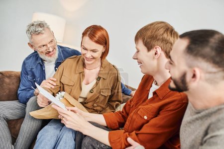 cheerful gay man showing wedding invitation to overjoyed parents in living room