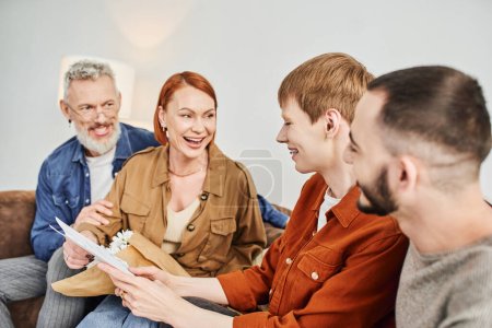 excited woman looking at son holding wedding invitation near gay partner at home