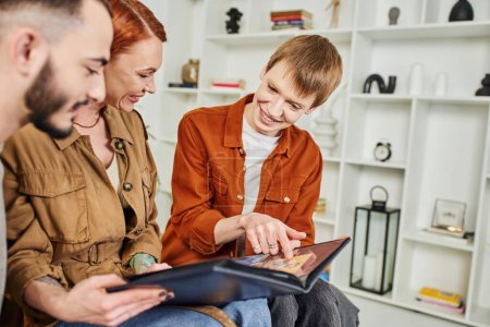 Photo for Smiling gay man pointing at photo in album near happy mother and blurred boyfriend at home - Royalty Free Image