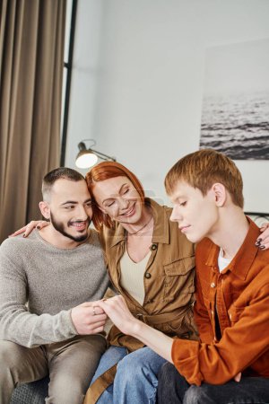 smiling woman embracing young gay man and happy son showing wedding ring at home