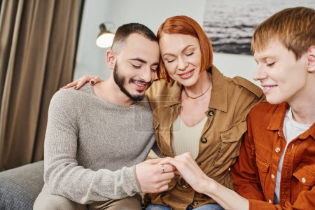 joyful gay man showing wedding ring to happy mother and gay partner at home