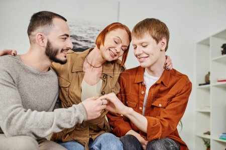 overjoyed woman with closed eyes embracing son with gay partner at home