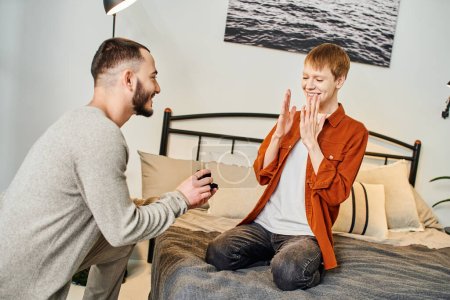 overjoyed redhead man showing wow gesture near gay partner holding jewelry box with wedding ring in bedroom