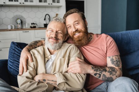 Fröhlich Homosexuell Mann umarmt bärtigen Partner während sitzen auf Couch zu Hause 