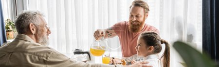 Smiling gay man pouring orange juice near daughter and partner at home, banner 