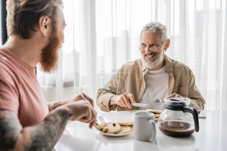 Cheerful gay man talking to blurred tattooed partner during breakfast at home 