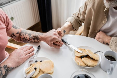 Recortado vista de tatuado pareja gay cogido de la mano cerca de panqueques y café en casa 