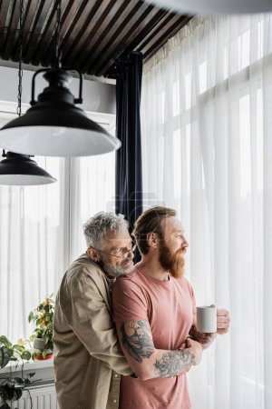 Mature gay man hugging tattooed partner with cup of coffee at home 