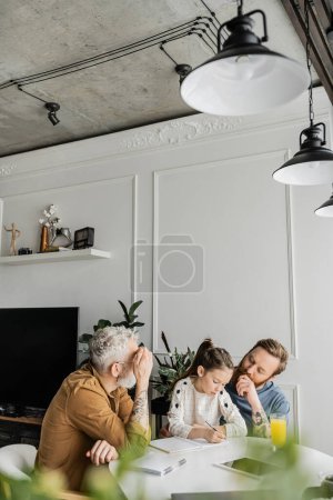 Chica preadolescente haciendo deberes con padres gay cerca de jugo de naranja y tableta digital en casa 