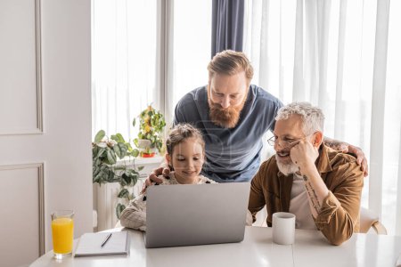 Smiling gay parents with daughter using laptop near notebook and drinks at home 