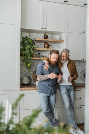 Smiling gay man holding cup and hugging partner with cellphone in kitchen 