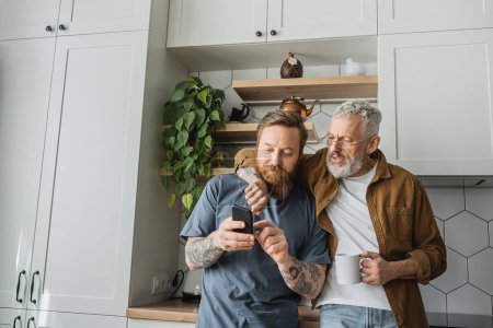 Schwuler Mann mit Tasse Kaffee umarmt Partner mit Smartphone in Küche 