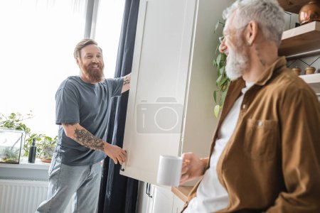Cheerful tattooed gay man looking at blurred partner with coffee near cupboard in kitchen 