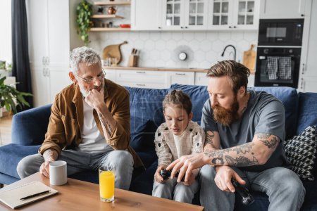 Photo for KYIV, UKRAINE - APRIL 19, 2023: Tattooed gay dad helping daughter with joystick near partner and drinks at home - Royalty Free Image