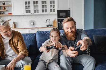 Photo for KYIV, UKRAINE - APRIL 19, 2023: Preteen girl playing video game near smiling homosexual dads at home - Royalty Free Image