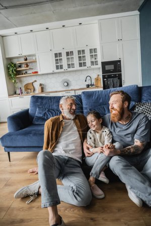 Alegres padres del mismo sexo sentados cerca de la hija adoptada en el suelo en casa 