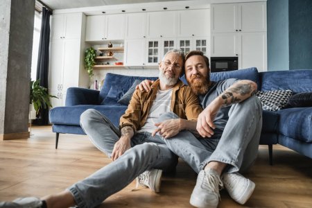 Lächelnde schwule Männer schauen in die Kamera, während sie zu Hause auf dem Boden sitzen 