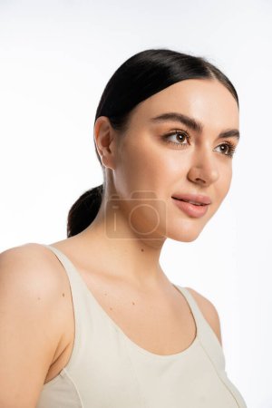 mujer hermosa y joven con cabello moreno, belleza natural y piel perfecta mirando hacia otro lado mientras posa en camiseta aislada sobre fondo blanco  