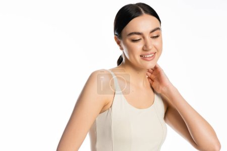 schöne junge Frau mit natürlichem Make-up, brünetten Haaren und perfekter Haut lächelnd, während sie im Tank-Top steht, wegschaut und mit der Hand am Hals isoliert auf weißem Hintergrund posiert