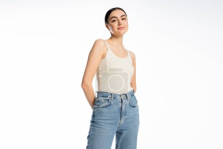 appealing and young woman with natural makeup, brunette hair and perfect skin smiling while posing in tank top and denim jeans and looking at camera on white background 