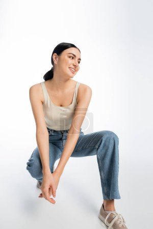 full length of lovely young woman with natural makeup, brunette hair and perfect skin smiling while sitting in denim jeans and tank top while looking away on white background 