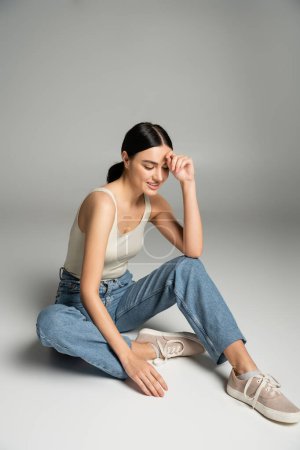 full length of charming young woman with natural makeup, brunette hair and perfect skin smiling while sitting in denim jeans and tank top while looking down on grey background 