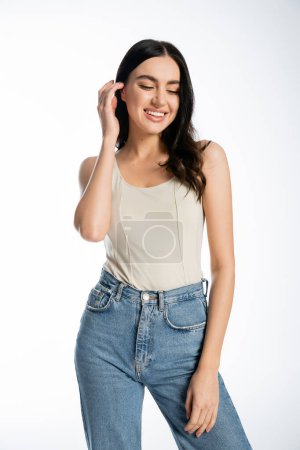 charming woman with natural makeup and perfect skin smiling, adjusting shiny brunette hair while posing in denim jeans and tank top and looking away on white background 
