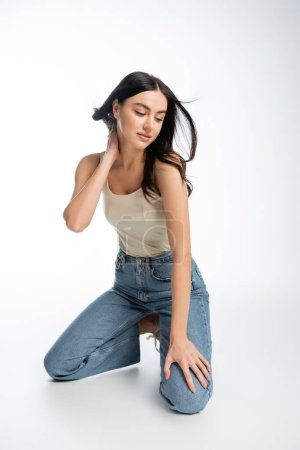 full length of young and alluring woman with natural makeup, brunette hair and perfect skin standing on knees and posing in denim jeans with tank top while looking away on white background 