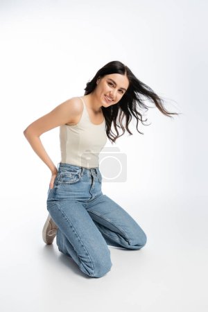full length of young and gorgeous woman with flawless makeup, brunette hair and perfect skin standing on knees and posing in denim jeans with tank top while on white background 