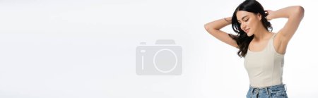 Foto de Mujer joven y seductora con maquillaje natural, cabello moreno y piel perfecta posando en camiseta con las manos detrás de la cabeza y sonriendo aislada sobre fondo blanco, pancarta - Imagen libre de derechos