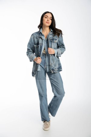 full length of alluring young model with brunette hair, flawless and natural makeup posing in denim jacket and blue jeans while standing and looking at camera on white background 