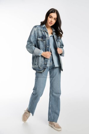 full length of charming young model with brunette hair, flawless and natural makeup posing in denim jacket and blue jeans while walking and looking at camera on white background 