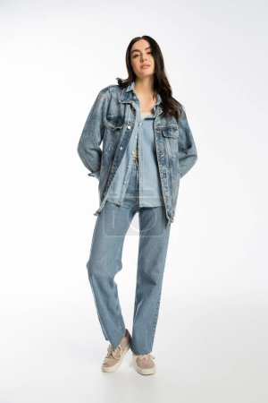 full length of fashionable young model with brunette hair and flawless makeup posing in denim jacket and blue jeans while standing and looking at camera on white background 