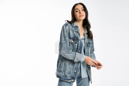 Photo for Fashionable young model with brunette hair and flawless makeup posing in blue denim jacket and jeans while standing and looking at camera on white background - Royalty Free Image