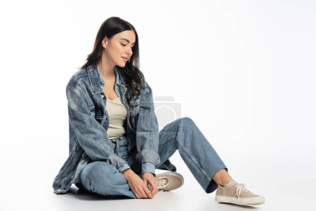 in voller Länge verführerische junge Frau mit natürlichem Make-up und brünetten Haaren posiert im trendigen Jeans-Outfit, während sie auf weißem Hintergrund sitzt und wegschaut
