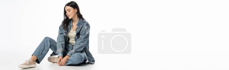 Téléchargez les photos : Pleine longueur de femme séduisante avec maquillage naturel et cheveux bruns posant en tenue denim à la mode tout en étant assis et en regardant loin sur fond blanc, bannière - en image libre de droit