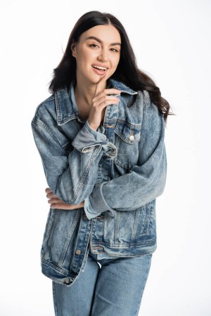 happy young woman with gorgeous brunette hair posing in stylish blue denim jacket and holding finger near face while looking at camera on white background
