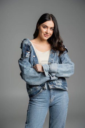 alluring young woman with gorgeous hair posing with folded arms, standing in blue jeans and denim jacket and smiling while looking at camera on grey background