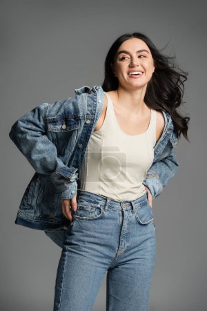 happy young woman with gorgeous brunette hair posing with hands on waist and standing in stylish blue jeans and denim jacket while looking away on grey background